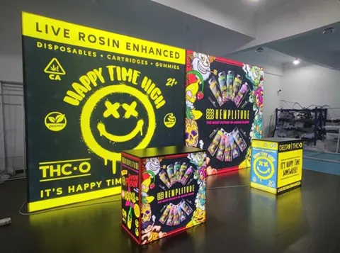 Happy Time High / Hemplitude trade show booth built by Purple Exhibits: Bold black and neon-yellow Happy Time High backlit wall with a smiley graphic, a graphic-wrapped Hemplitude counter and a small cube lightbox, set u