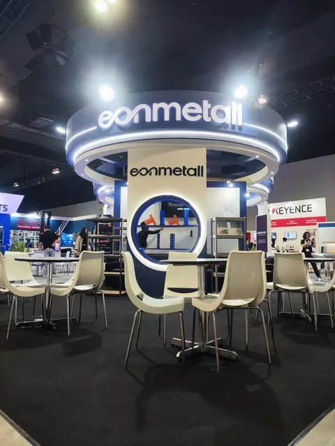 Eonmetall trade show booth built by Purple Exhibits: Portrait view of the Eonmetall island booth showing the ring-lit circular header with white logo, a porthole showcase of shelving hardware, and white chairs at round t