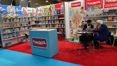 Funskool trade show booth built by Purple Exhibits: Funskool toy booth with colourful wall shelving packed with games and toys, a light-blue branded counter on red carpet and two men in meeting at a table beside Play & L