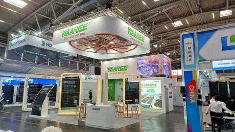 Waaree trade show booth built by Purple Exhibits: Waaree solar island booth at a European show with a large overhead truss ring and green logo header, a circular orange wheel graphic, LED screen, backlit module spec pane