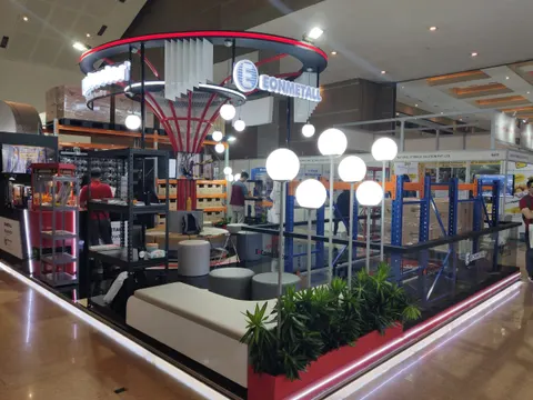 Eonmetall trade show booth built by Purple Exhibits: Eonmetall island booth with a circular red and black overhead ring, a red tree-like column structure, white globe pendant lights, dark grey lounge seating with planter