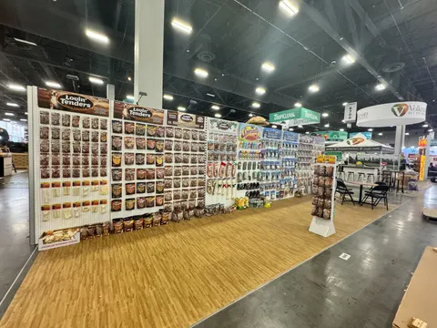 Louis Tenders trade show booth built by Purple Exhibits: Wide angle of a long inline booth for Louis Tenders with white slatwall panels hung with chicken tender and snack packaging, brown header signs, a spinner rack and