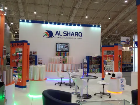 Al Sharq trade show booth built by Purple Exhibits: Al Sharq Flexible Packaging Factories white back wall with orange and blue product towers, film-roll displays lit by green LED, a wall screen and glass bar tables with 