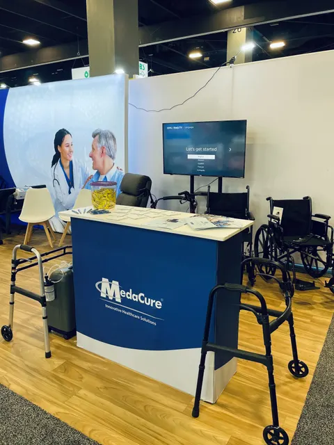 MedaCure trade show booth built by Purple Exhibits: MedaCure inline booth with a large white and blue fabric backwall of a doctor and patient, blue branded counter, wall-mounted TV, wood-look flooring and wheelchairs and
