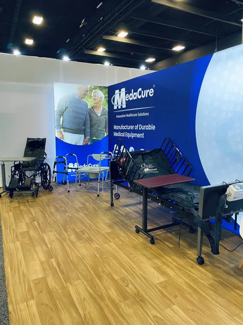 MedaCure trade show booth built by Purple Exhibits: MedaCure 'Manufacturer of Durable Medical Equipment' blue and white backwall with a senior couple graphic, a hospital bed, wheelchairs and walkers on wood-look flooring