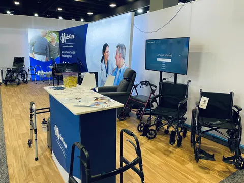 MedaCure trade show booth built by Purple Exhibits: Wide view of the MedaCure inline booth: long white and blue fabric backwall, blue counter with brochures, TV screen and a row of wheelchairs and rollators on wood-look