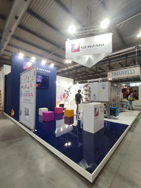 Senzani trade show booth built by Purple Exhibits: Senzani island booth with a glossy navy-blue raised floor with white edging, white hanging sign, blue backwall with Omnia graphic, colourful cube seats, a '70' anniversa