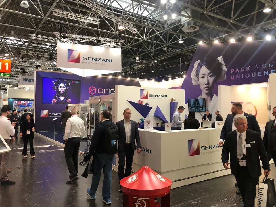 Senzani booth by Purple Exhibits: busy show-floor view of the white Senzani island booth with a hanging sign