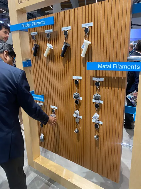 Trade show booth built by Purple Exhibits: Close-up of a slatted oak product wall with hooks holding 3D-printing filament samples under blue 'Flexible Filaments' and 'Metal Filaments' labels, with a visitor handling one.