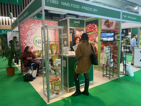 NIMS Food trade show booth built by Purple Exhibits: Small NIMS Food freeze-dry inline booth in dark green with pink berry graphics, a glass product showcase, a white counter and staff and a visitor in the foreground on