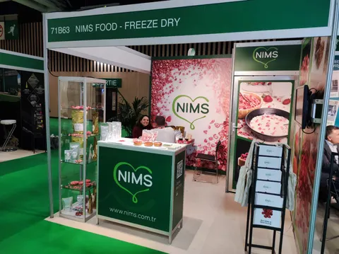 NIMS Food trade show booth built by Purple Exhibits: NIMS Food inline booth with dark green NIMS logo counter, glass showcase tower, pink berry graphic back wall and a staff member behind the counter on green carpet.