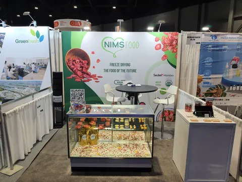 NIMS Food trade show booth built by Purple Exhibits: Frontal view of the NIMS Food freeze-drying inline booth with a berry graphic back wall, a glass display case of freeze-dried products, a round bar table with white st