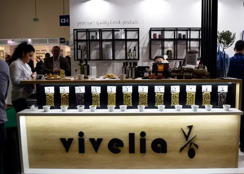 Vivelia trade show booth built by Purple Exhibits: Close-up of a light wood Vivelia counter with a row of glass jars of olives and tasting cups, black shelving with Greek products behind, an olive tree and staff and visi