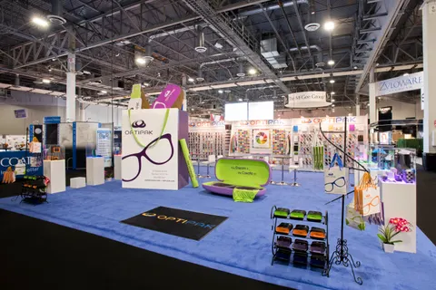 Optipak trade show booth built by Purple Exhibits: Clean wide shot of the Optipak island booth with blue carpet, white pedestals, oversized purple glasses graphics and green eyewear-case props with metal display racks.