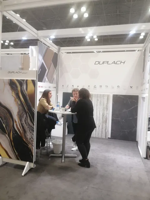 Duplach trade show booth built by Purple Exhibits: White Duplach booth showing large-format porcelain slabs in gold-veined marble, dark marble and wood-look finishes on the walls, with staff and a visitor talking at a ro