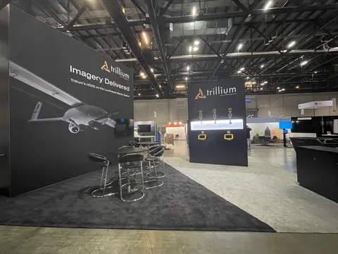 Trillium trade show booth built by Purple Exhibits: Wide view of the Trillium black island booth showing the tall drone graphic wall, black bar-height tables and stools, a black tower with backlit gimbal camera display a