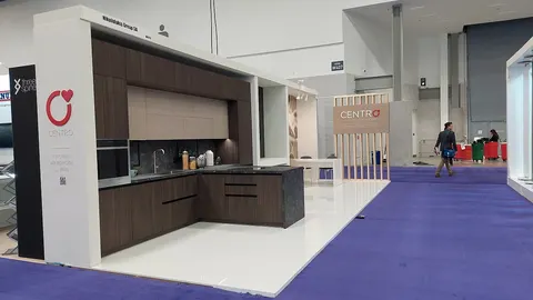 Centro trade show booth built by Purple Exhibits: Centro kitchen showroom booth with white structural frame, dark walnut cabinetry, glossy white floor and a wood slat divider wall, seen from the aisle over purple carpet.