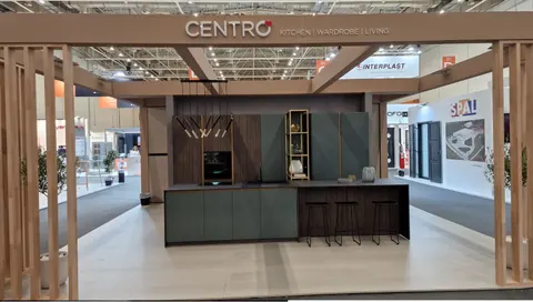 Centro trade show booth built by Purple Exhibits: Centro 'Kitchen Wardrobe Living' booth framed by timber slat portal, displaying a dark green and walnut kitchen with island, black bar stools, brass shelving and pendant