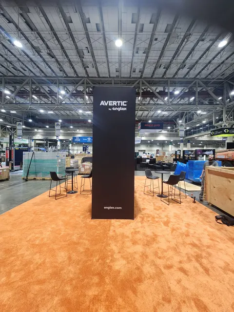 Avertic by Engtex trade show booth built by Purple Exhibits: Tall black branded tower reading Avertic by Engtex and engtex.com standing on bright orange carpet with bistro tables and stools on either side; show floor sti
