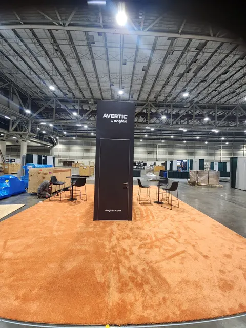 Avertic by Engtex trade show booth built by Purple Exhibits: Rear view of the Avertic by Engtex black tower showing its hidden storage door, on orange carpet with bar-height tables and chairs; crates and setup crews visi