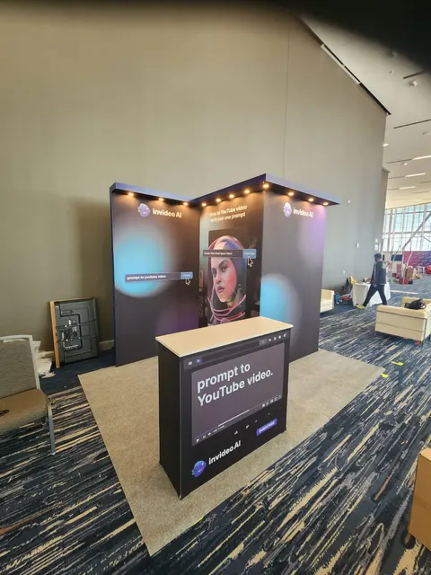 invideo AI trade show booth built by Purple Exhibits: Dark purple-toned angled fabric back wall for invideo AI with app-interface graphics and overhead spotlights, fronted by a branded counter reading prompt to YouTube v