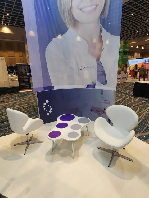 SpecialtyRx trade show booth built by Purple Exhibits: Close-up of a SpecialtyRx lounge nook with two white swan chairs and a purple-dot coffee table on white raised flooring beneath a large fabric graphic of a smiling p