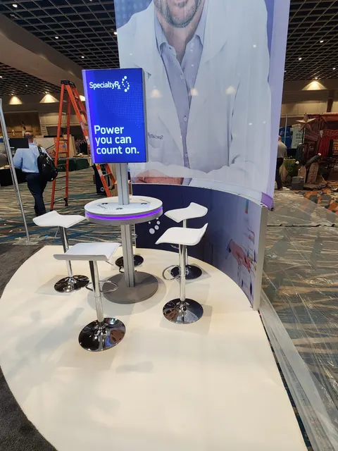 SpecialtyRx trade show booth built by Purple Exhibits: Detail of a SpecialtyRx bar-height round table with white stools and a blue lightbox reading Power you can count on, white flooring, with a pharmacist graphic tower