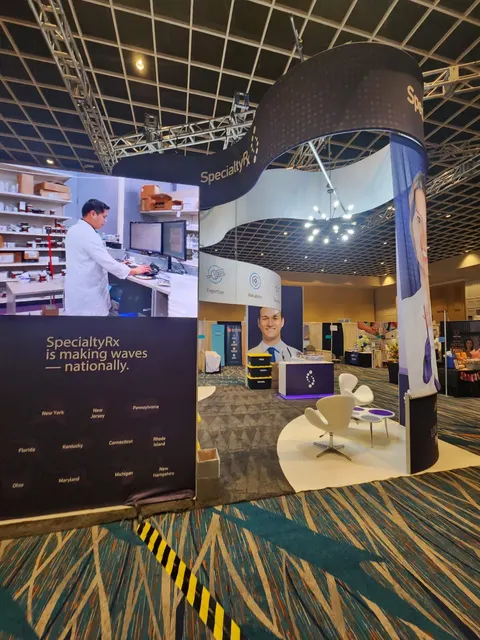 SpecialtyRx trade show booth built by Purple Exhibits: Angled view of the SpecialtyRx island showing a navy wall graphic reading SpecialtyRx is making waves nationally with a state map, curved overhead header, pharmacist