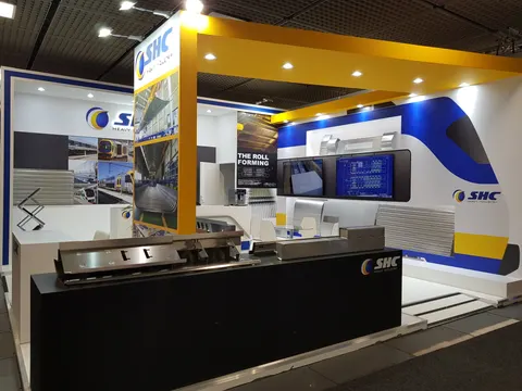 SHC Heavy Industry trade show booth built by Purple Exhibits: SHC Heavy Industry booth in white, yellow and navy with a yellow overhead canopy, a black reception counter with metal roll-formed samples, train-graphic back