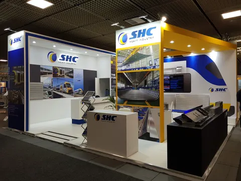 SHC Heavy Industry trade show booth built by Purple Exhibits: Wide view of the SHC Heavy Industry booth showing the yellow header, navy end wall, factory photo panels, white counter with logo, black sample display and a