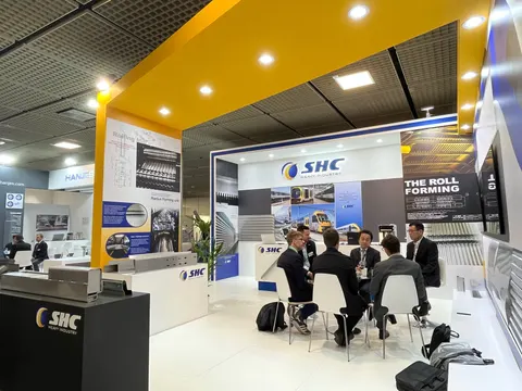 SHC Heavy Industry trade show booth built by Purple Exhibits: SHC Heavy Industry booth in use with a group of visitors seated at a meeting table under the yellow canopy, roll-forming graphics and metal profile samples on