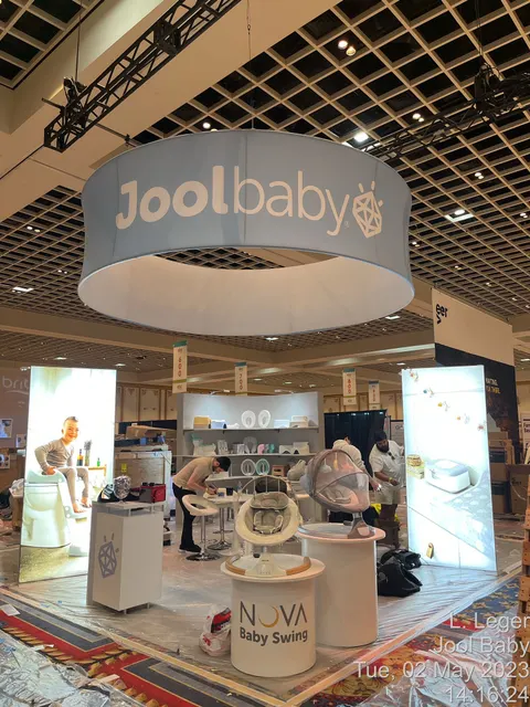 Jool Baby trade show booth built by Purple Exhibits: Jool Baby island booth mid-setup with a large light-blue circular hanging sign, backlit lifestyle graphic towers, white slatwall product bays and round NOVA Baby Swing
