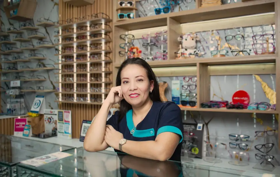 Smiling optician leaning on a glass counter in front of wooden shelves full of eyeglass frames