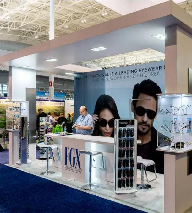 FGX International eyewear booth by Purple Exhibits with a white canopy, a lifestyle print of people in sunglasses, frame towers, a lit glass case and a counter with stools