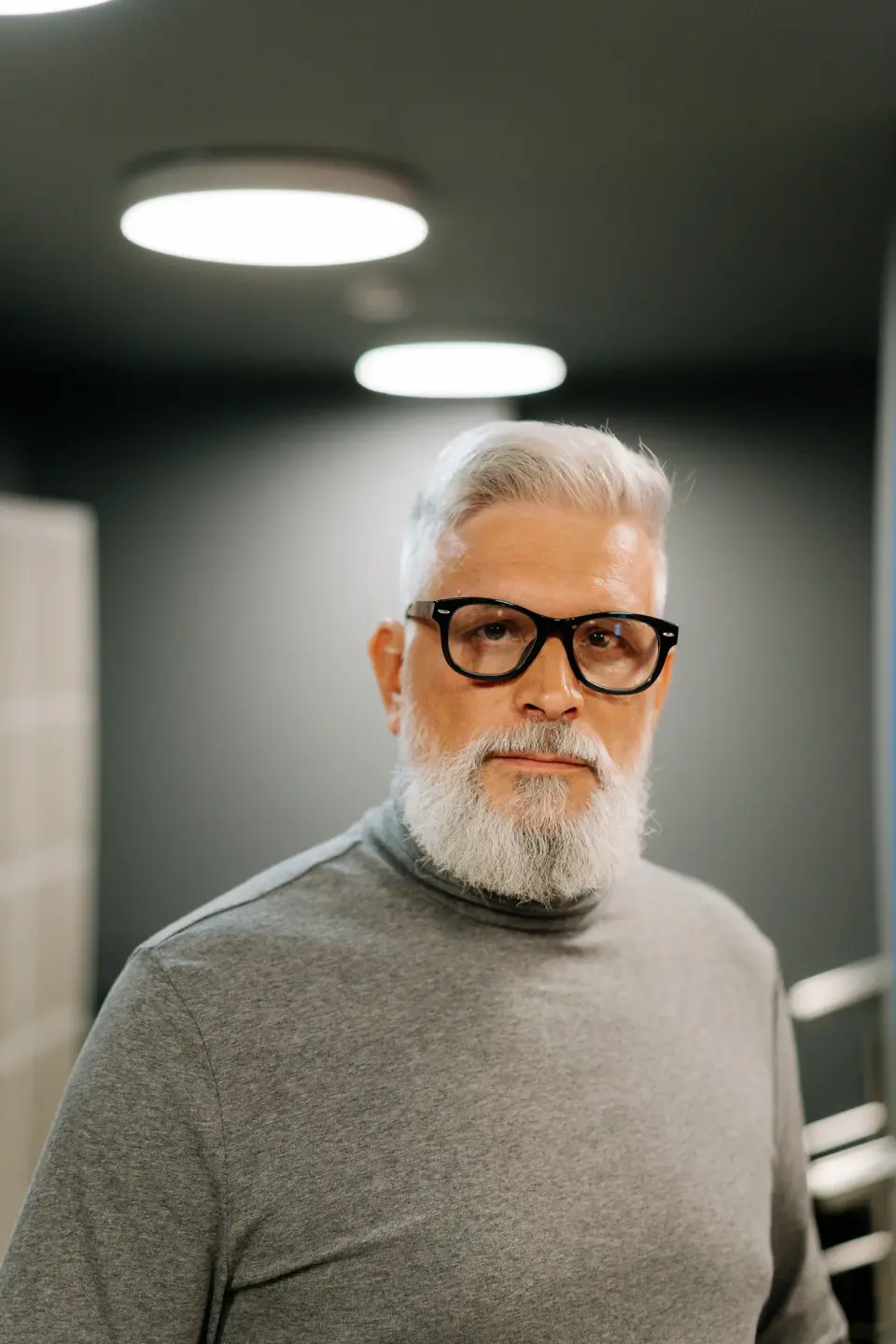 Silver-haired bearded man in bold black eyeglass frames and a gray turtleneck