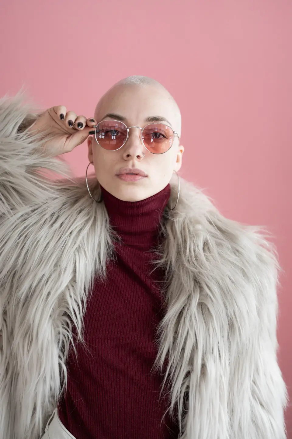 Woman in round metal sunglasses with pink lenses and a faux fur coat on a pink backdrop
