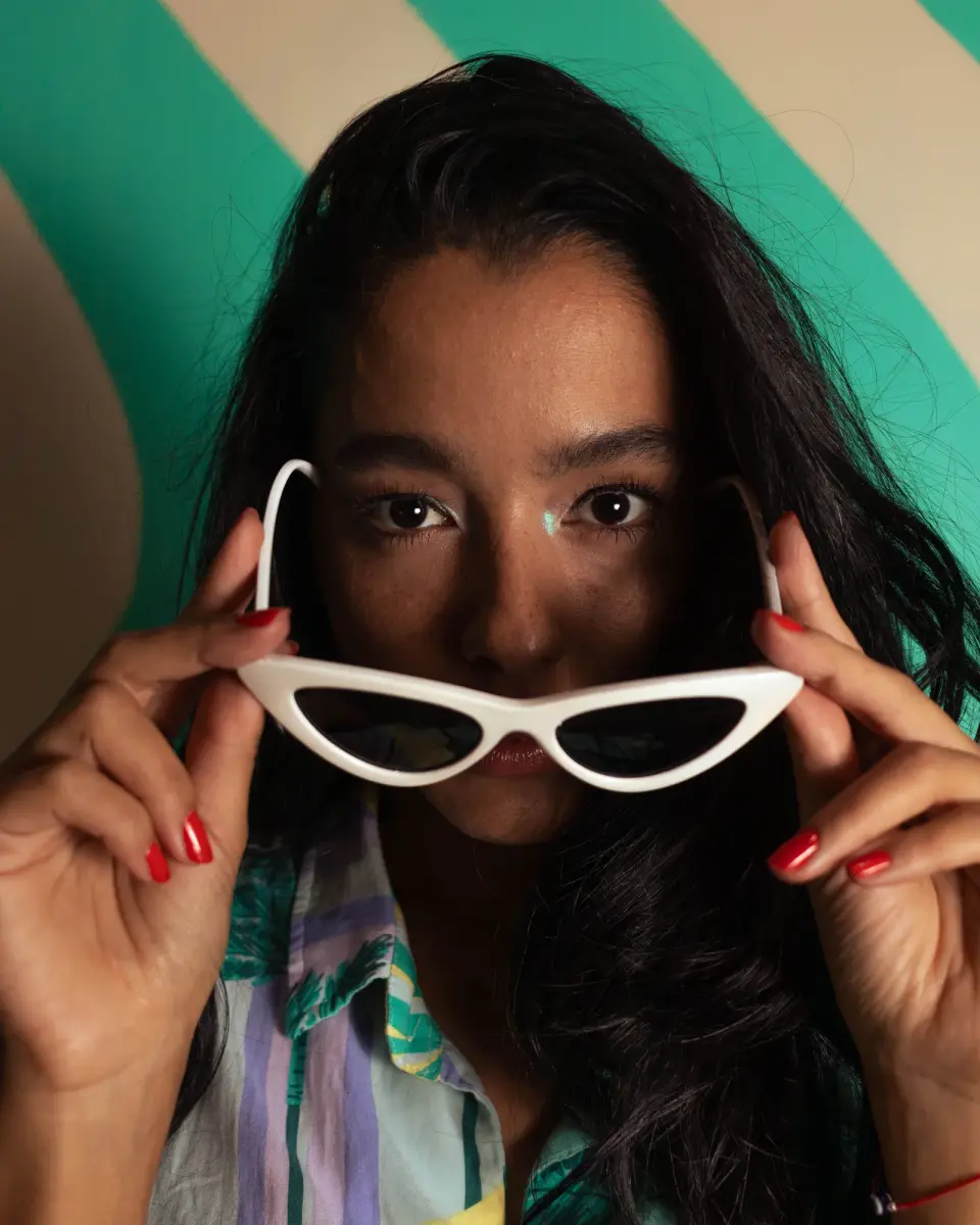 Woman holding white retro cat-eye sunglasses below her eyes, colorful studio portrait