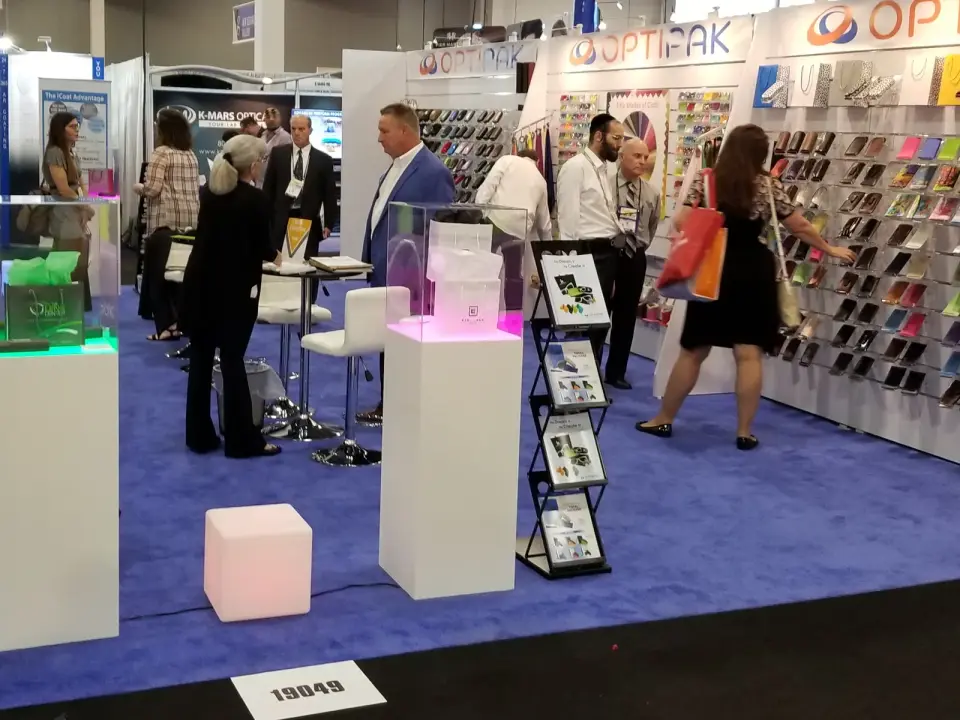 Buyers walking the Optipak booth by Purple Exhibits past lit white pedestals and a wall of colored eyewear cases