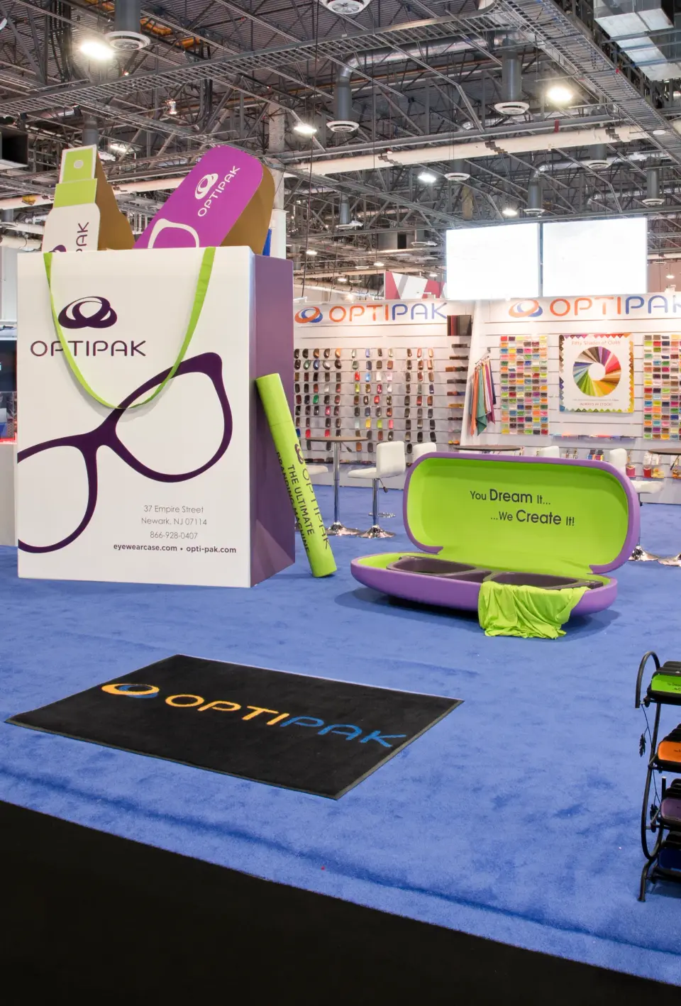 Optipak eyewear case booth by Purple Exhibits with a giant printed shopping bag, an oversized open glasses case used as a bench and a blue floor