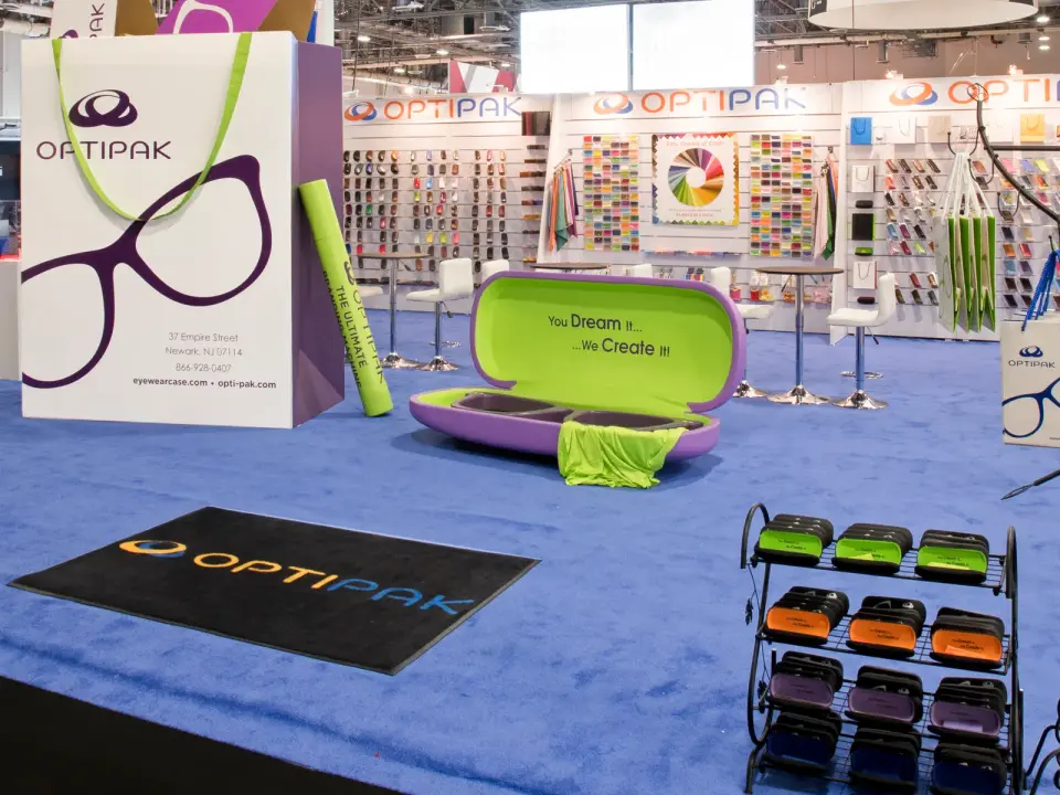 Close view of the Optipak booth by Purple Exhibits with a giant printed bag, an oversized glasses case bench and stacked case displays on blue carpet