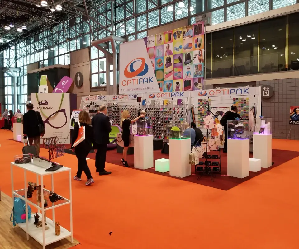 Optipak island booth by Purple Exhibits with lit white pedestals under clear covers, a sample wall and a tall branded banner on an orange aisle