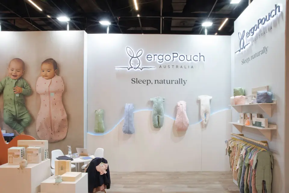 ergoPouch booth by Purple Exhibits with a lit logo wall, baby sleep suits hung along a lit curve, a large print of two babies, a garment rail and shelves of boxed product