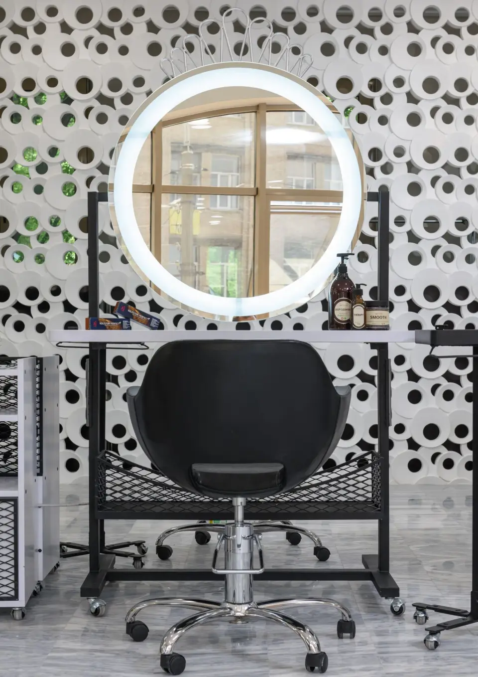 A salon styling station with a round ring-light mirror, a black chair and a patterned wall