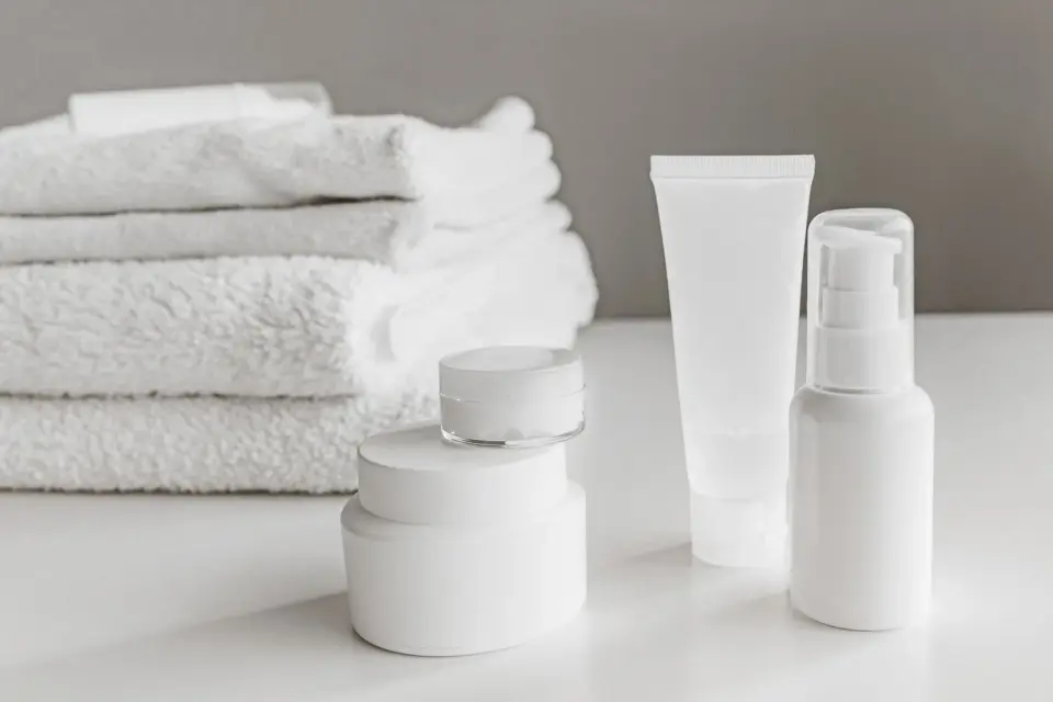 White skincare jars, a pump bottle and a tube beside rolled white towels