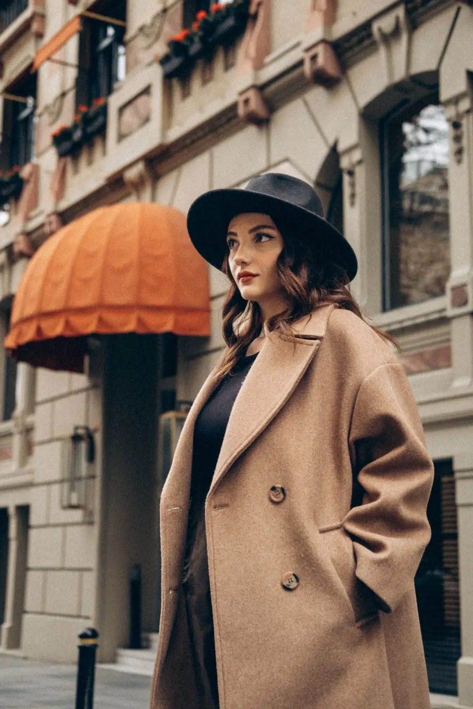 Woman in an oversized camel coat and black felt hat on a city street, street style portrait