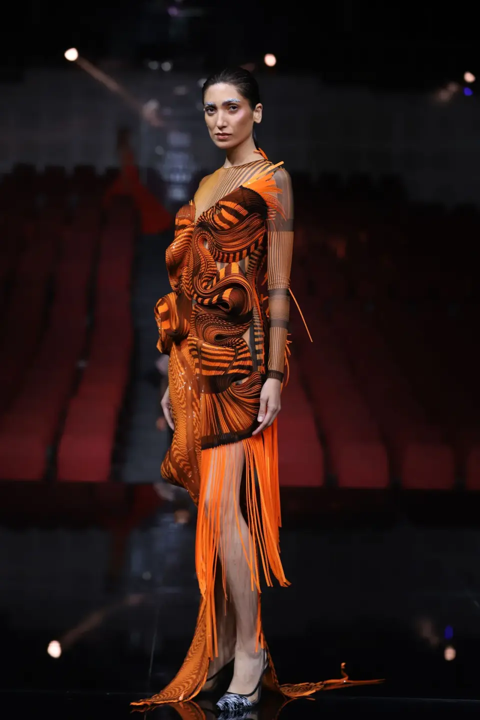 Model on a runway in a sculpted orange and brown couture dress with fringe