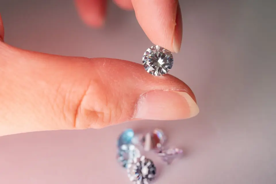 Fingers hold a round brilliant diamond above a scatter of loose colored stones