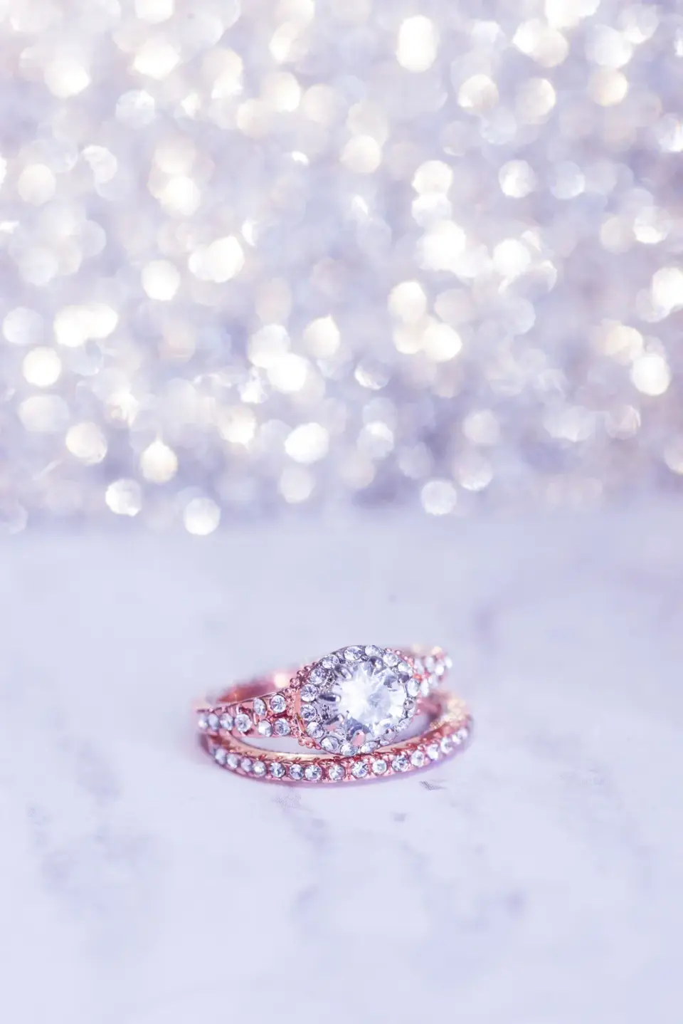 A rose gold diamond engagement ring and band on white marble with soft sparkle behind