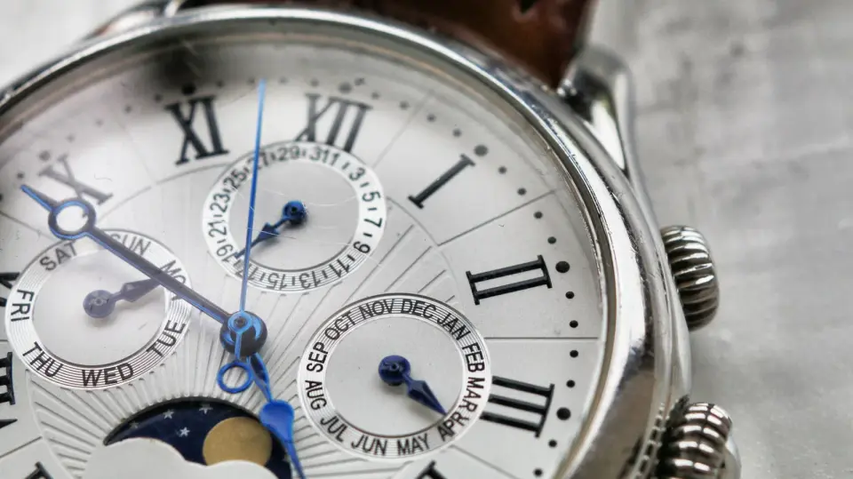 Close view of a steel watch with Roman numerals, blue hands, date, day and month dials and a moon phase