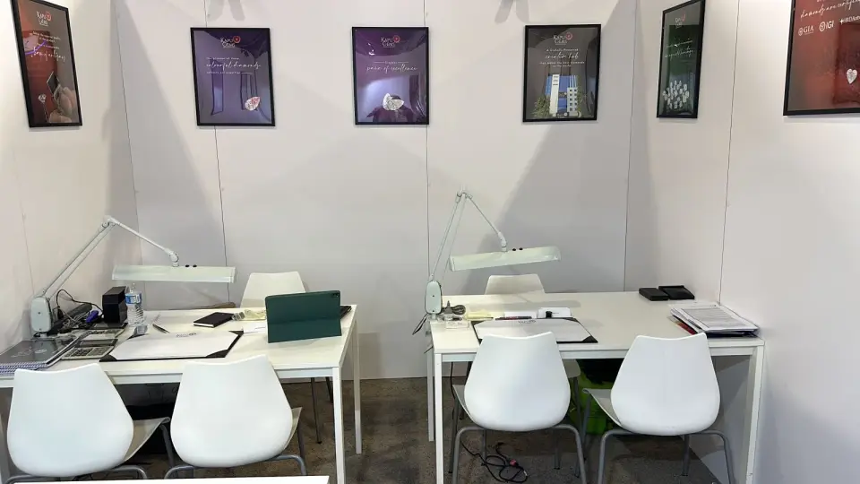 Private buying room in the Kapu Gems booth by Purple Exhibits with two white work tables, daylight task lamps, white chairs and framed diamond prints